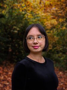 Julie Lew, a half-Chinese/half-white person with short, dark brown hair, stands outdoors in a park with fall foliage, wearing a black velvet dress and looking slightly off camera with a slight smile.