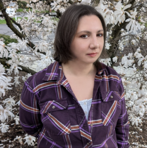 Justine Lombardi, a white woman with mid-length dark hair, sits outdoors under a dogwood tree in spring, and has a slight smile.