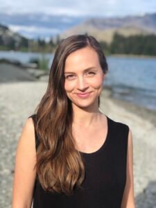 A headshot of author Kahli Scott, a white woman with long brown hair wearing a black short-sleeved jumpsuit. She is in focus, smiling with a closed mouth, and the blurred backdrop depicts a lake, mountains and trees.