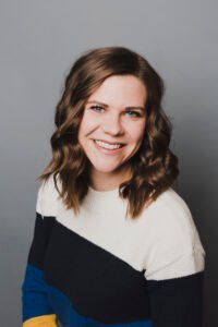 Portrait of a white woman with shoulder-length brown hair. She is posed against a gray background and is smiling at the camera.