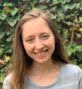 Karen Myna Cantor, a white woman with long brown hair, stands in front of a wall of ivy with her head tilted and a smile on her face.