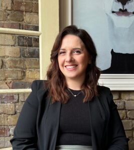 A woman with shoulder-length brown hair smiles while sitting indoors against a brick wall with string lights and a modern portrait painting in the background. She is wearing a black blazer over a black top and a delicate necklace.