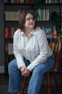 Kat Stoddard, a white woman with shoulder length brown hair and glasses, sits hunched slightly forward with her hands clasped. She's wearing jeans and a pinstriped button-down and looking away from the camera. There are shelves full of books and plants behind her.