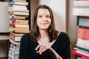A white woman with reddish-brown hair in a dark blue V-neck dress stares at the camera with a neutral expression. She is middle aged. One hand rests on her knees. She is wearing a silver ring. She is between two thin stacks of books, one in soft focus.