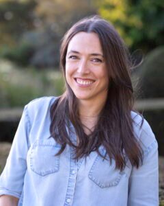 Kate Eberle stands outside in front of a backdrop of greenery. She is wearing a light blue chambray button-down shirt, her slightly windswept brown hair is down over her shoulders, and she is smiling at the camera.