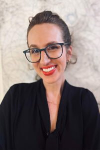 Kate Fox, a white woman with glasses and dark hair in a bun, smiles at the viewer.