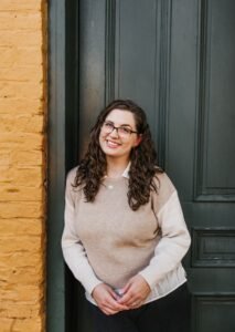 Kate Westbury, a white woman with brown wavy hair and glasses, stands against a green door and smiles at viewer.