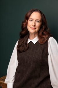 Kathleen Carr Foster, a white woman with long brown hair, leans against a wooden table. She is looking at the viewer and has a faint smile. The image has a forest green background.