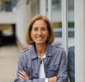 Kathleen Reidy, a white woman with brown, bobbed hair. She's wearing a jacket and blouse, and she is standing outside with her arms crossed. She's smiling.