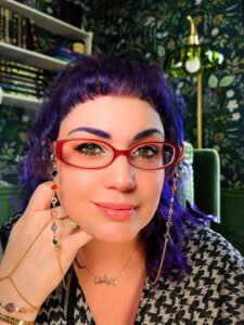 A light-skinned woman wearing a keffiyeh smiles at the camera, leaning her cheek on her hand. She has purple hair with micro bangs, and wears red glasses with a jeweled chain, a hand chain, and a gold necklace that says "كايتلين" or "Kaitlyn" in Arabic.