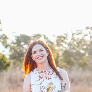 Katie Bowman, a white woman with long burgundy hair, standing outside. She is looking at the viewer and smiling.