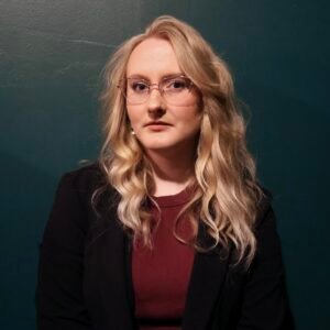 Katie Ortiz, a white woman with blonde hair, wear a black blazer, looking at viewers without smiling. In front of dark green wall.