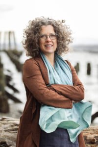 Julie Salmon Kelleher, a white woman with curly hair and glasses, stands outside with her arms crossed in the wind.