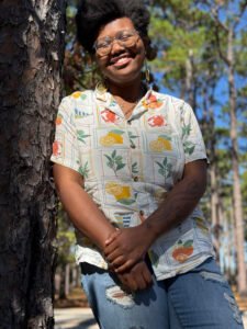 Kennedy Cole, a Black woman with glasses and short Afro, leans against a tree with her hands crossed in front of her waist. She wears glasses and a button up shirt, smiling.