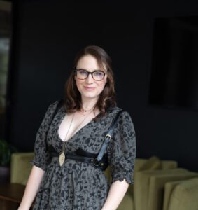 Kimberly Lynn Hanson, a white woman with brunette hair wearing a moss green dress with black accents, stands in front of a green couch and black background.