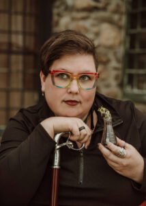 Kit Gray, a.k.a. Elise Scott, is a round-faced white nonbinary femme with short dark hair and thick-framed rainbow glasses. They sit before an old stone castle, gazing at the viewer. In their left hand is a small Erlenmeyer flask with an ivy vine growing out of it. In their right hand is a rosewood walking cane with a silver handle in the form of twisted vines.