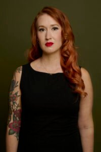 Krista Diamond, a white woman with long red hair and tattoos is standing against an olive green background wearing a black dress and looking pensively at the camera. The image is shot from the waist up.