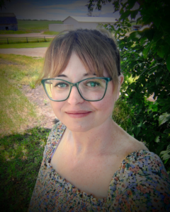 Lacey Moone, white woman with light brown hair and bangs wearing green eyeglasses and a floral print dress.