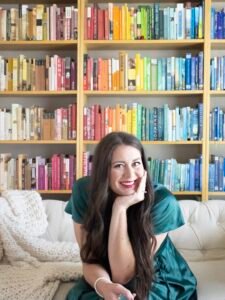 Laura Vogt, a white woman with long brown hair, sits in front of floor to ceiling rainbow bookshelves. She is looking at the viewer with a smile.