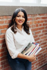 Lavanya Lakshmi, an Indian woman with black, wavy hair, stands against a brick wall, holding books and smiling at the camera.