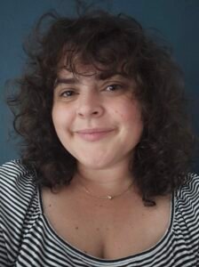 Leslie René, a white woman with curly dark brown hair, stands in front of a green wall wearing a black and white striped shirt.
