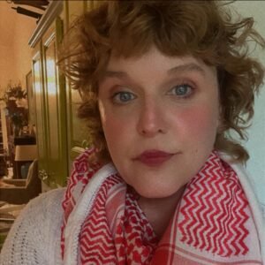 Libby Edwardson, a white woman with short hair that is wavy and light brown, and blue eyes wears a red and white keffiyeh scarf around her neck. She looks at the camera with a neutral expression.