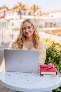Linda Hamilton, a white woman with blonde hair sitting at a table wearing a yellow dress. She has a laptop open in front of her, smiling at the viewer.