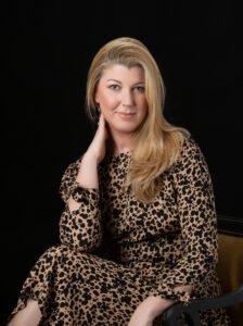 A woman with long blonde hair wearing a leopard-print dress sits on a chair against a black background, resting her hand on her neck and smiling at the camera.
