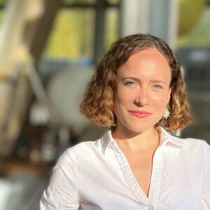 Author headshot of Lior Torenberg. She is smiling softly, with shoulder-length wavy light brown hair, wearing a white collared shirt and statement earrings. She stands in natural light with a softly blurred background of windows and greenery, creating a warm and inviting atmosphere.
