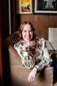 Liz Edelbrock, a white woman with short reddish blonde hair, sits in a chair resting her chin on one hand and smiling.