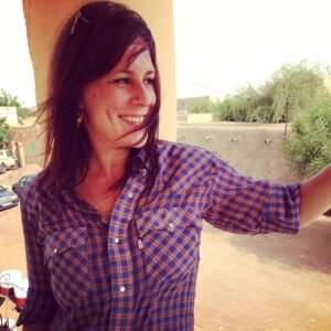 Liz Wanic, a white woman with long brown hair is standing in a tower with a concrete wall behind her in a desert location. The wind is blowing through her hair and she is looking to the side with a smile on her face.