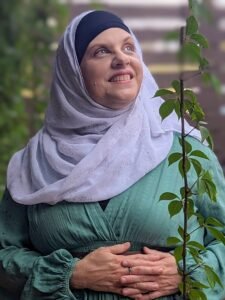 Loretta is wearing a light green dress and a white hijab and is surrounded by hanging vines. She is looking upward with a mysterious expression.