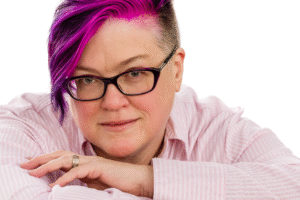 M.J. Beasi, a white, nonbinary person wearing glasses, with pink/purple/blue undercut hairstyle, leans on their arms in a close-up shot. They are looking at the viewer and have a slight smile.