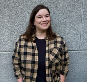 Maria Millage, a white woman with brown hair stands against a gray wall facing the camera. She is looking at the viewer and smiling.