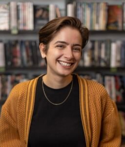 Marissa Macy, a white woman with short brown hair, wearing a yellow cardigan. She is smiling in front of a shelf full of movies.