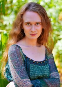 Marley Rose-Teter, a white woman with long golden hair, wears a green dress in front of a backdrop of foliage. She has her arms crossed and an enigmatic smile.