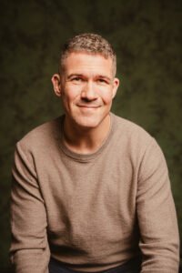 Marshall J. Moore is a white man with short graying brown hair smiling gently at the camera. He wears a beige shirt against a green backdrop.