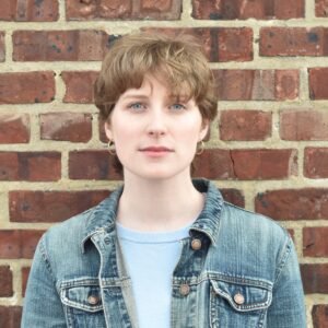 Mary Berman, a white woman with short brown hair, stands against a brick wall in a denim jacket and looks at the viewer.