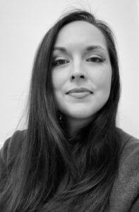 Mattea Orr, a black and white photo of a white woman with long dark hair wearing a gray sweater against a pale background. She is smiling at the viewer.