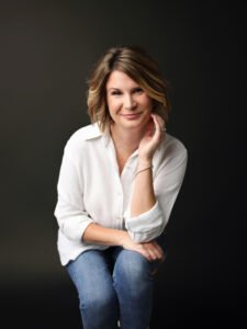 Meg Anderson, A woman leaning forward on a stool with her hand resting against the side of her face, wearing a white button up shirt and jeans.