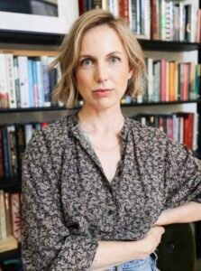Meghan Nesmith, a white woman with short blonde hair, sits in front of a bookshelf. She is looking at the viewer and has a slight smile.