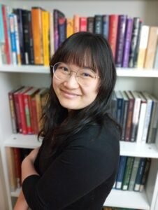 Mengxi Seeley, a Chinese American woman with long black hair, wearing round glasses and a black T-shirt, stands in front of a colorful bookshelf with her arms crossed. She is smiling at the viewer.