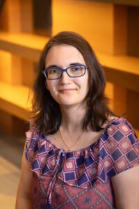 Millie Abecassis, a white woman with long brown hair, brown eyes, wearing blue glasses and a pink and purple t-shirt. She is smiling at the camera.