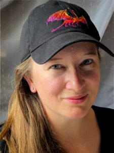 Mir Benitz, a white woman with long blonde hair and wearing a black cap with a vibrant rainbow dragon, smiles into the camera.
