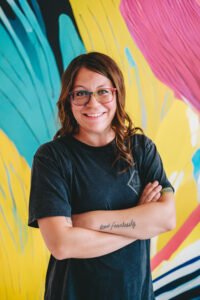 Mira Gonzalez, a white woman with long curled dark hair stands in front of a bright yellow, pink, and tourquoise wall with her arms crossed. She is wearing a black tee shirt and rainbow glasses and is smiling at the viewer.