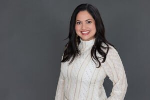 Author photo for Mona Tewari showing a smiling woman with long, dark hair who is wearing a white top with vertical gold accents and diagonal mirrored accents. SHe is wearing chunky white and gold earrings and has red lipstick.