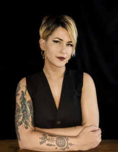 Natalie Kikić, a white woman was brown hair and blonde highlights, poses with her arms resting on a table. She is wearing a black vest and has tattoos on her arms. The background is black.