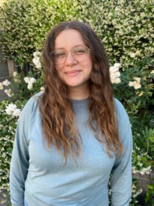 Natalie Messier, a woman with pale skin and wavy brown hair, stands in front of a jasmine bush, smiling at the viewer.