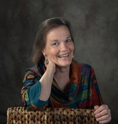 Nicola Ranson leans on the back of a wicker chair supporting her head with her right arm. She smiles warmly and the light catches her eyes.