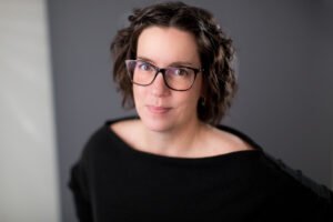 Nicole Brooks, a white woman with curly brown hair and glasses, stands with a black boat neck shirt, is looking at the viewer with a slight smile.
